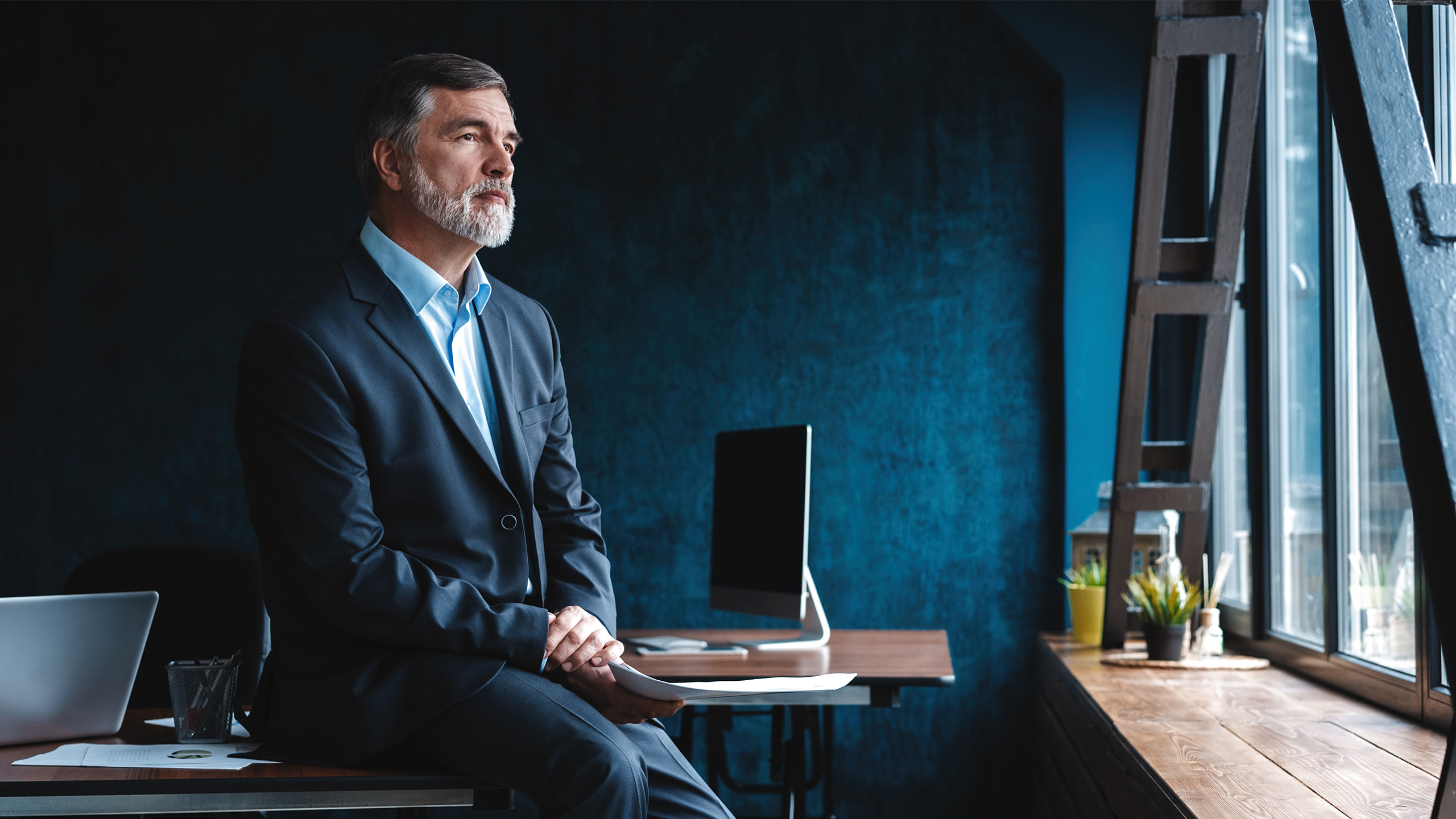 Person dressed in business attire sitting on the edge of a desk in a dimly lit office, with a computer monitor and workspace items visible in the background.